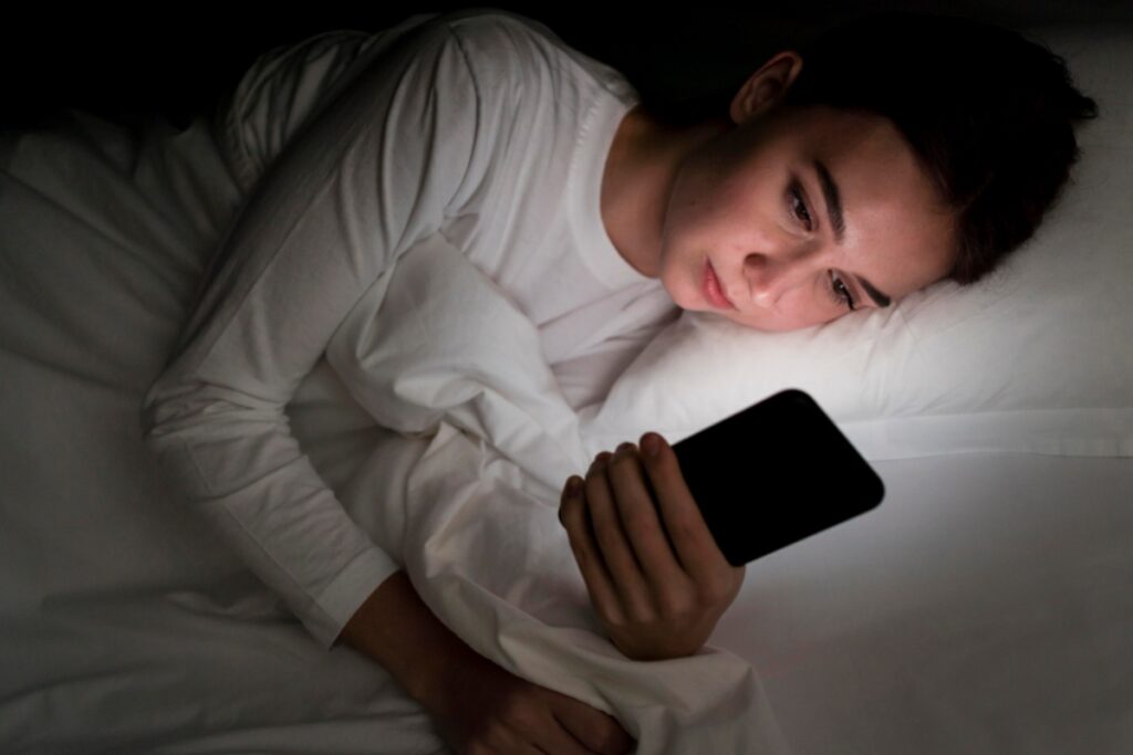 Žena ležící v posteli osvětlená displejem telefonu.